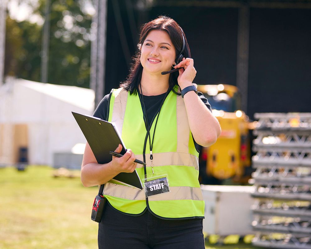 Eine Eventmanagerin koordiniert eine Outdoor-Veranstaltung über ihr Headset. Sie trägt eine Warnweste und hält ein Klemmbrett in der Hand.