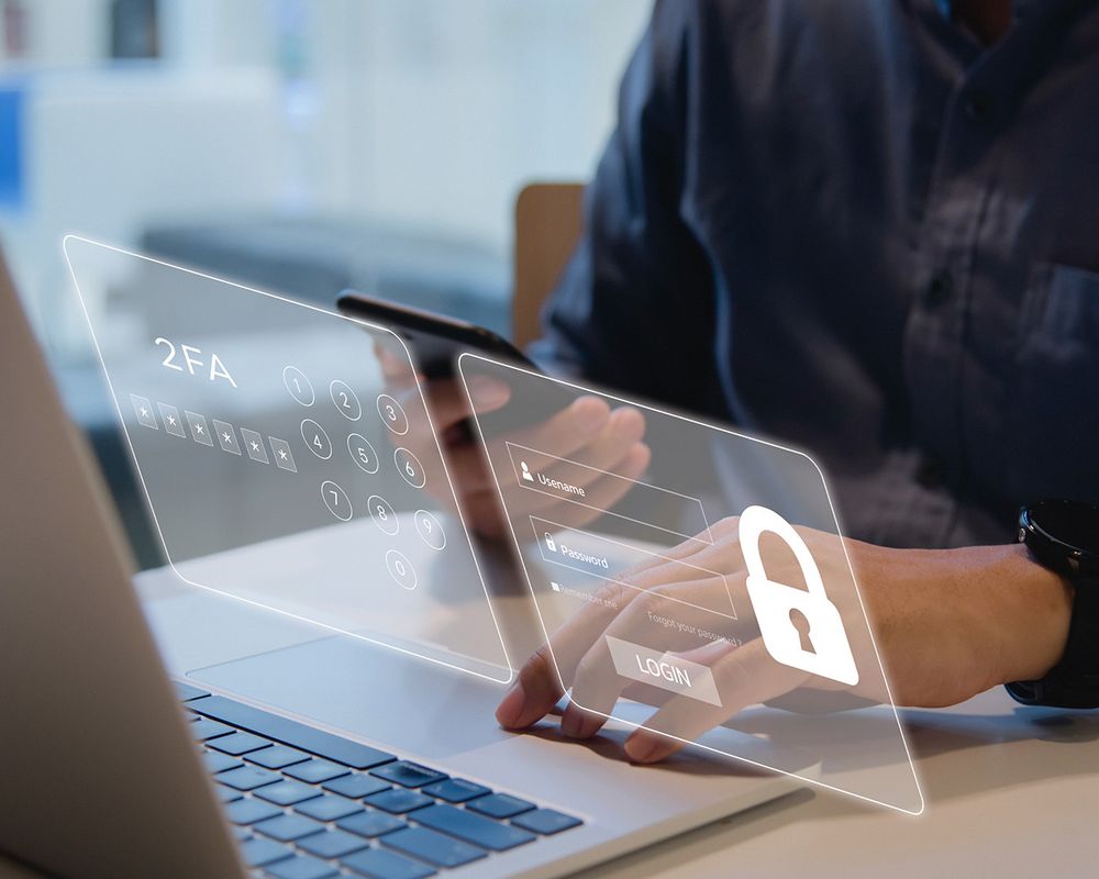 Ein Mann sitzt an einem Schreibtisch und tippt auf seinem Laptop. Über dem Laptop ist eine Fläche mit einem digitalen Login angedeutet, der mit einer 2FA geschützt ist.