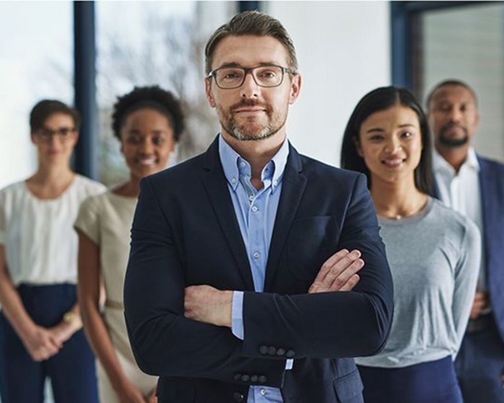 Eine Führungskraft steht selbstbewusst der Kamera zugewendet. Hinter ihm stehen mehrere seiner Mitarbeitenden.