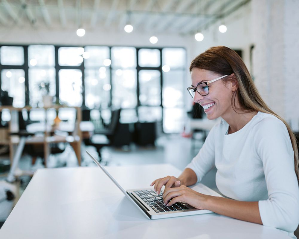 Eine Mitarbeiterin sitzt mit ihrem Laptop am Schreibtisch und lächelt.