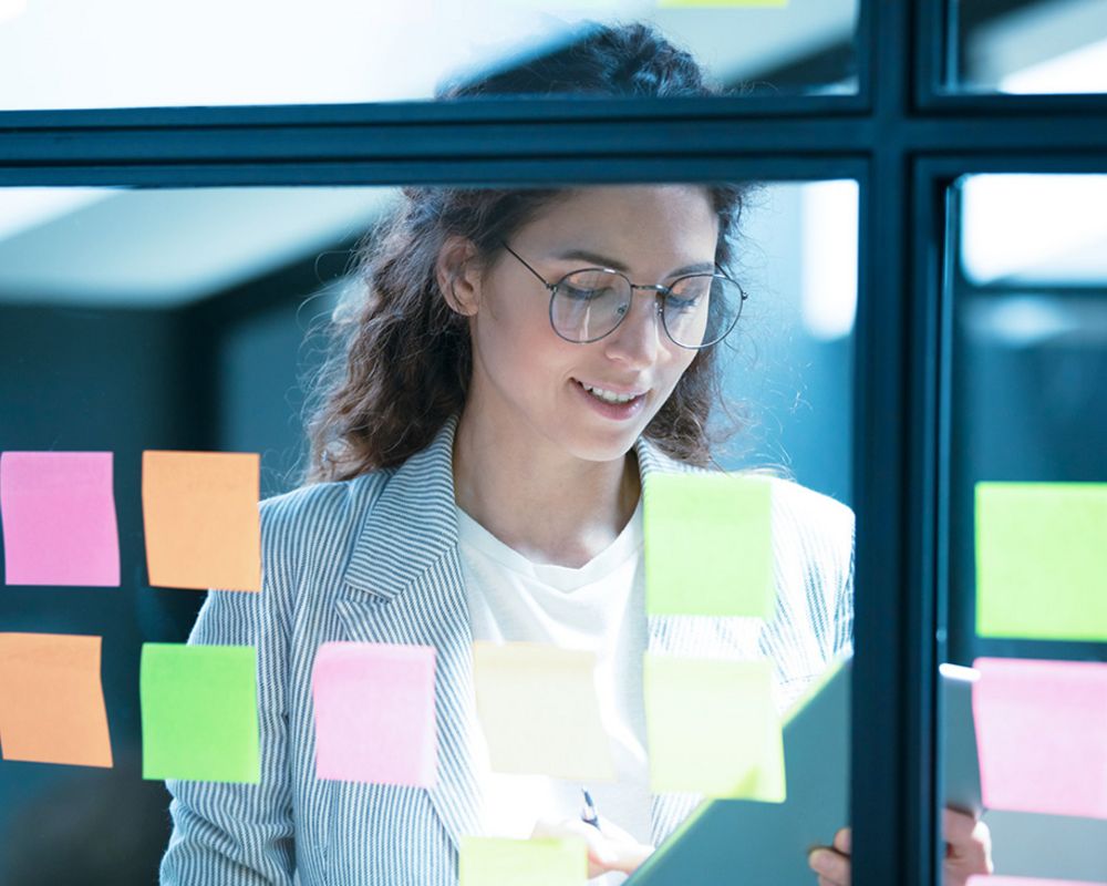 Eine Projektmitarbeiterin geht einen Projektablauf auf ihrem Tablet durch. Vor ihr an den Glaswänden hängen bunte Post its.