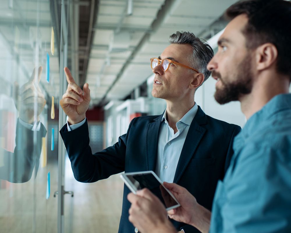 Zwei Kollegen besprechen das Leistungsangebot und betrachten eine Übersicht aus Post its an der Wand.
