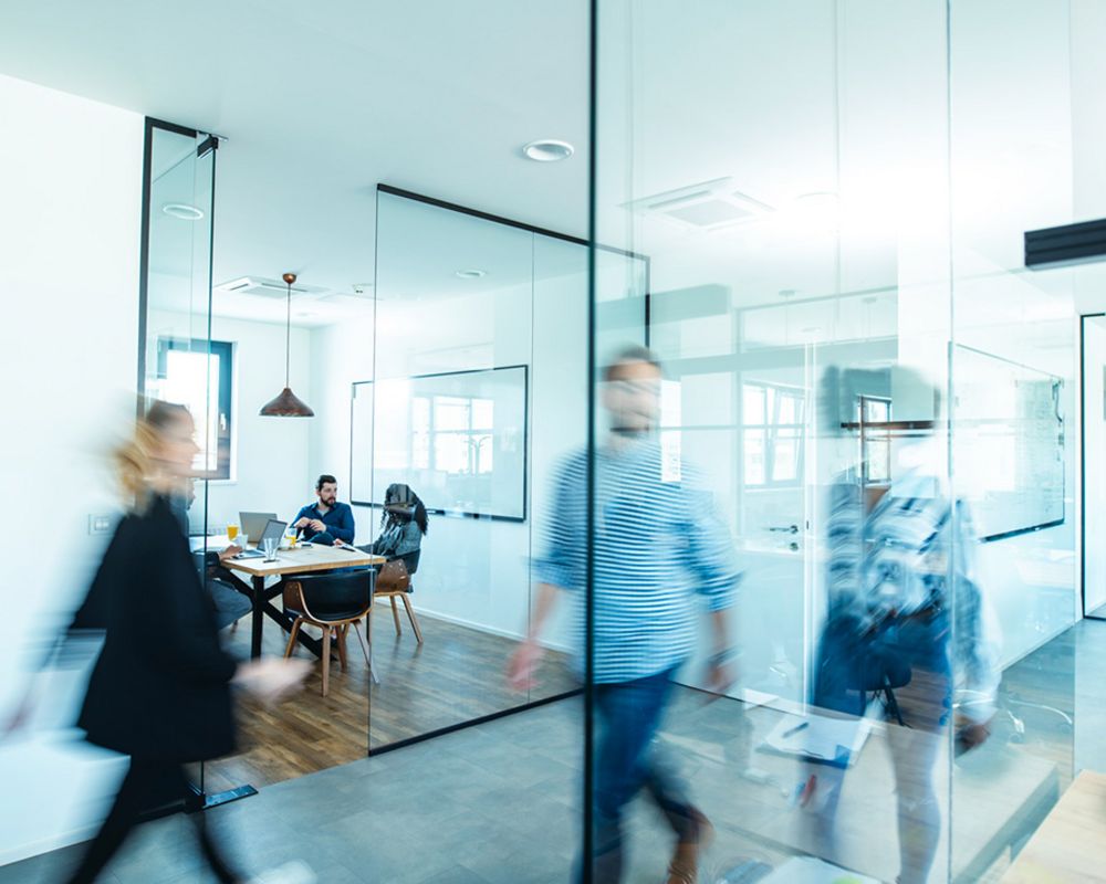 Im Hintergrund ist in einem Büro ein Beratungsgespräch zu erkennen. Im Vordergrund sind verschwommene Menschen in Bewegung zu sehen, die den Büroflur entlang laufen.